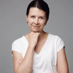 Portret uśmiechniętej kobiety z krótkimi, ciemnymi włosami, opierającej brodę na dłoni, z zegarkiem na ręce i złotym łańcuszkiem na szyi, na szarym tle.