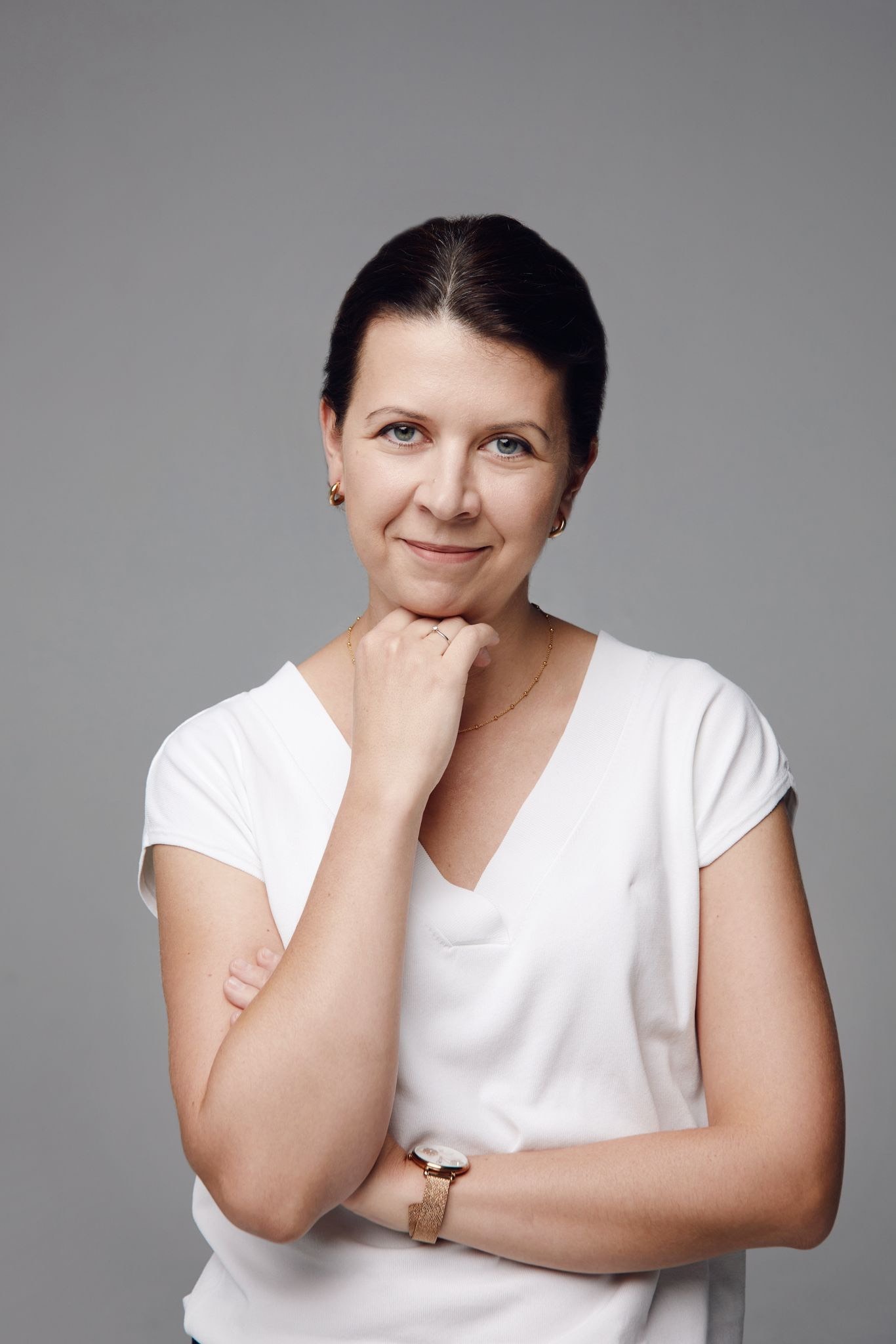 Portret uśmiechniętej kobiety z krótkimi, ciemnymi włosami, opierającej brodę na dłoni, z zegarkiem na ręce i złotym łańcuszkiem na szyi, na szarym tle.