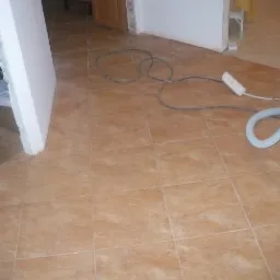 Układanie brązowych płytek podłogowych w trakcie prac wykończeniowych, widoczne przewody elektryczne, wąż od odkurzacza i fragment płyty gipsowej o białej powierzchni.