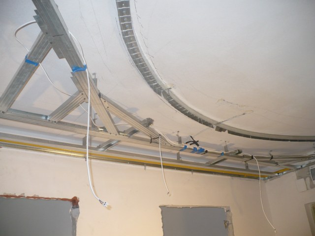 Metalowa konstrukcja sufitu podwieszanego, widoczne profile i kable, surowy stan wnętrza z fragmentami niezabezpieczonych ścian.