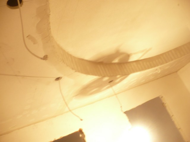 Niedokończony sufit z widocznymi przewodami elektrycznymi i fragmentami szpachlowania, oświetlony ciepłym światłem, ukazujący etap prac wykończeniowych.