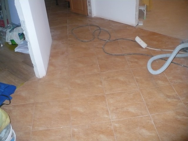 Układanie brązowych płytek podłogowych w trakcie prac wykończeniowych, widoczne przewody elektryczne, wąż od odkurzacza i fragment płyty gipsowej o białej powierzchni.