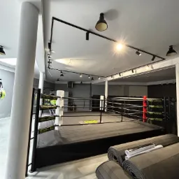 Nowoczesna sala treningowa z ringiem bokserskim o czarno-białej kolorystyce, oświetlona reflektorami sufitowymi, z matami do ćwiczeń zwiniętymi w rolki przy ringu.