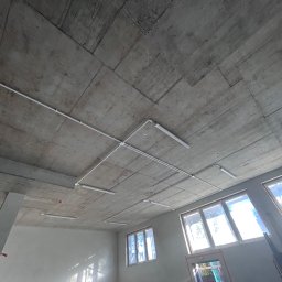 PerLOCK Patryk Sielawa - Surowy betonowy sufit z widocznymi przewodami i lampami fluorescencyjnymi, wnętrze w trakcie wykończenia, okna w tle.