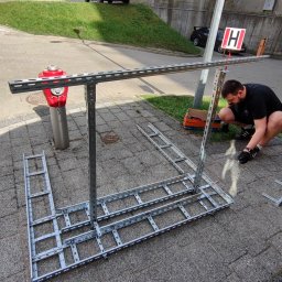 PerLOCK Patryk Sielawa - Pracownik w Białymstoku montuje metalową konstrukcję szkieletową na chodniku z kostki, obok czerwonego hydrantu i skrzynki narzędziowej Bosch, z użyciem wkrętarki akumulatorowej.