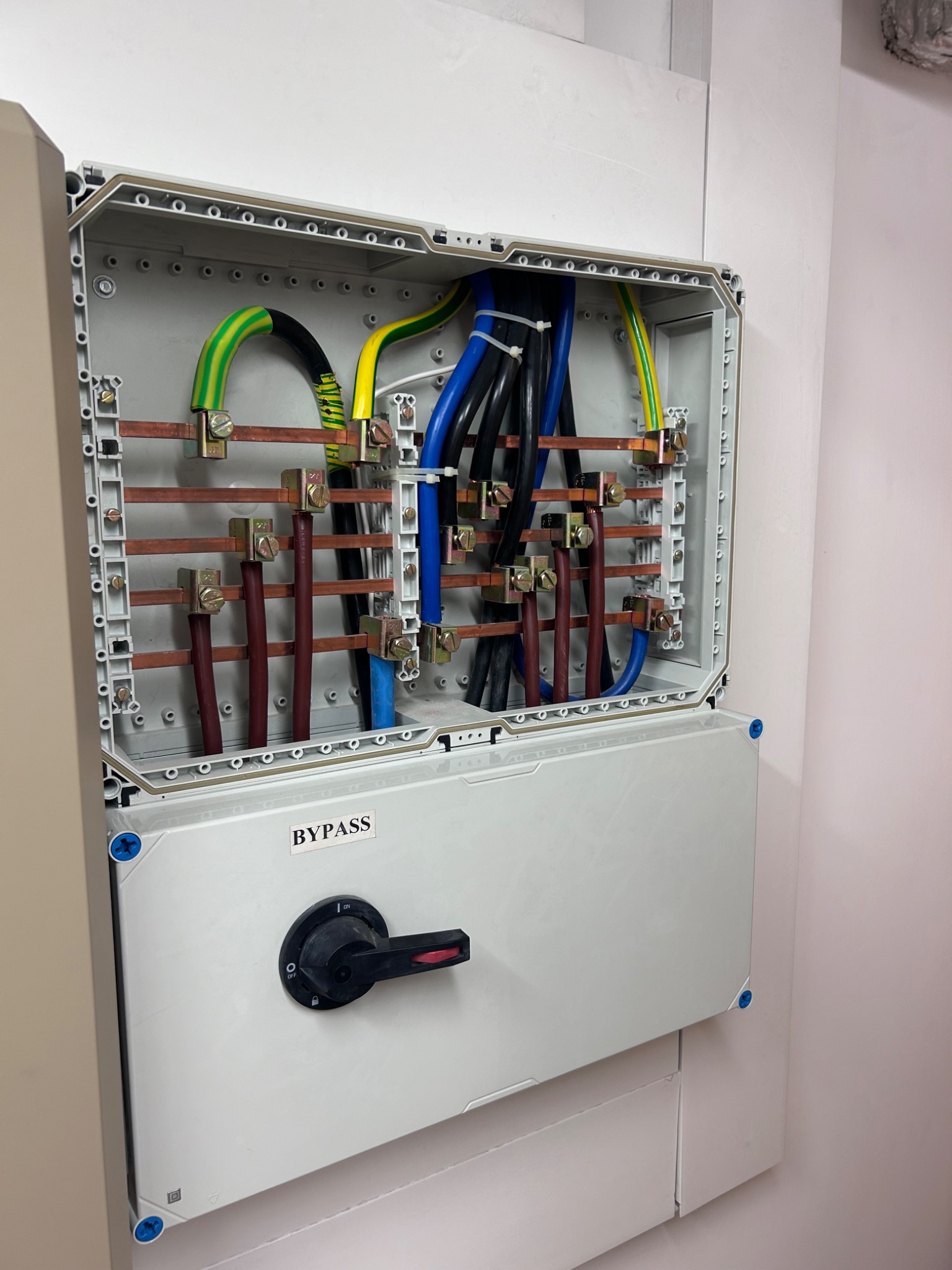 Otwarta skrzynka elektryczna z widocznymi przewodami w kolorach zielono-żółtym, niebieskim i czarnym, połączonymi z miedzianymi szynami. Na obudowie napis 'BYPASS' i przełącznik w pozycji OFF.