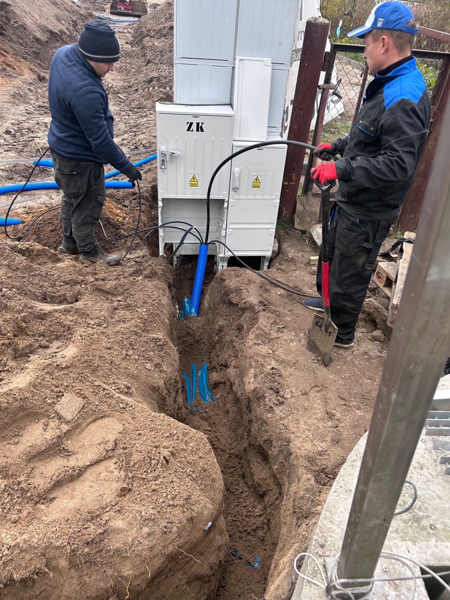 Dwóch pracowników w trakcie podłączania kabli do białej skrzynki elektrycznej ZK umieszczonej nad wykopem z niebieskimi rurami karbowanymi
