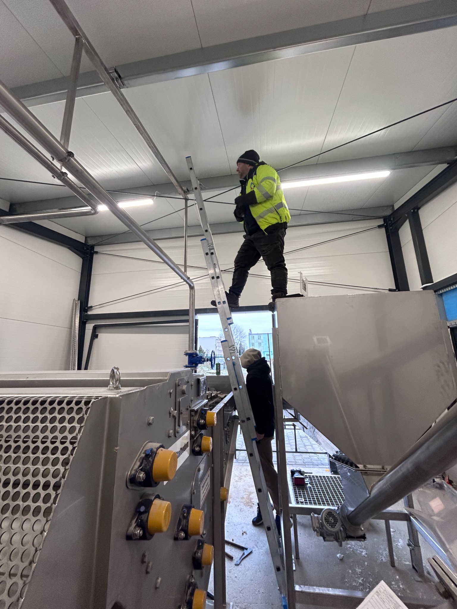 Elektryk w żółtej kurtce na drabinie montuje oświetlenie w hali produkcyjnej z nową maszyną przemysłową.