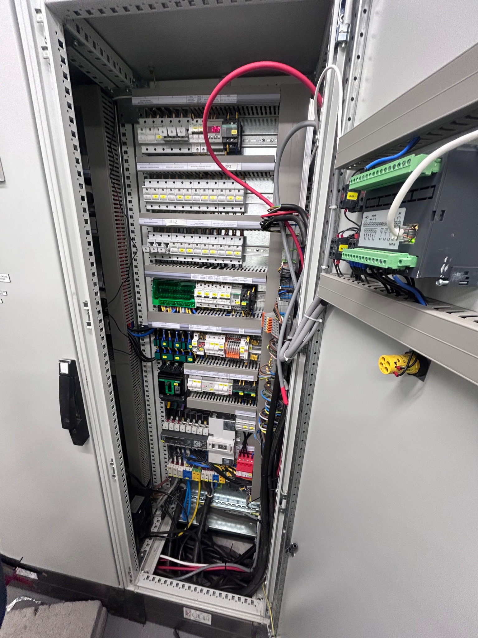 Otwarta szafa sterownicza z widocznymi bezpiecznikami, przewodami i modułami elektronicznymi, uporządkowanymi na szynach montażowych.