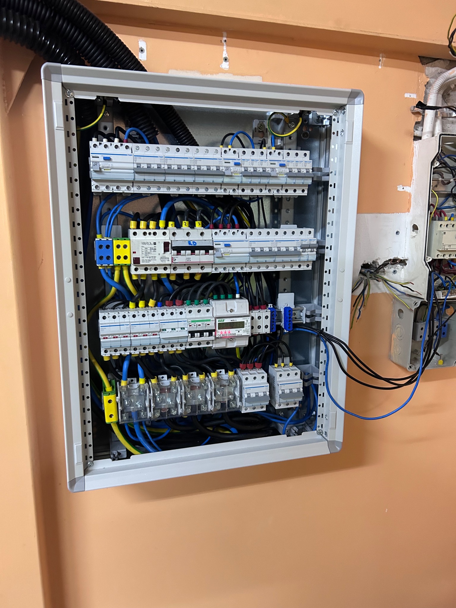 Otwarta skrzynka rozdzielcza z bezpiecznikami, okablowaniem w kolorach niebieskim, żółtym i czarnym, zamontowana na pomarańczowej ścianie. Widoczne elementy instalacji elektrycznej obok.