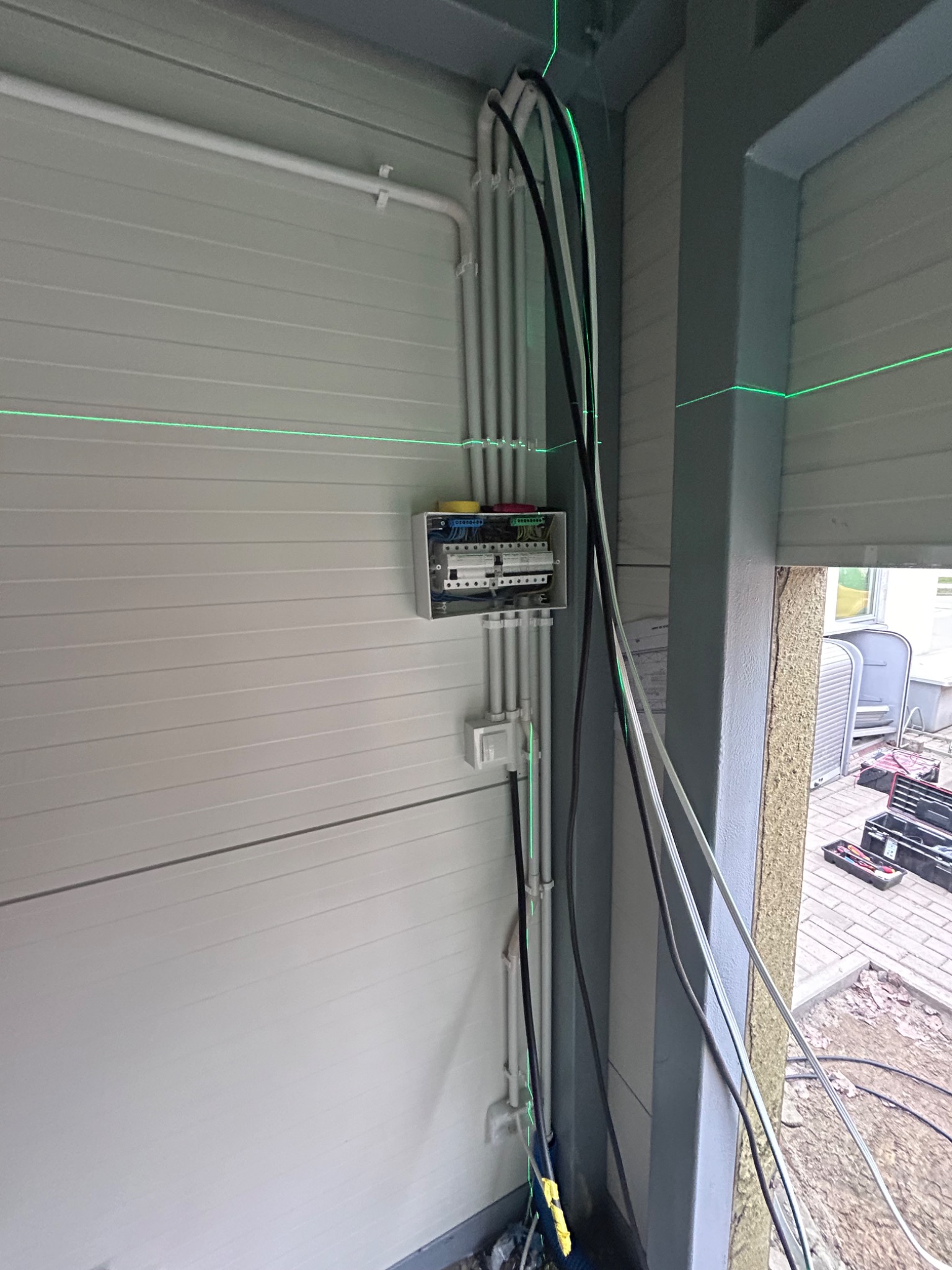 Szara ściana z zamontowaną skrzynką elektryczną, widoczne kable w peszlach, użycie lasera krzyżowego do poziomowania, narzędzia i skrzynka narzędziowa w tle.