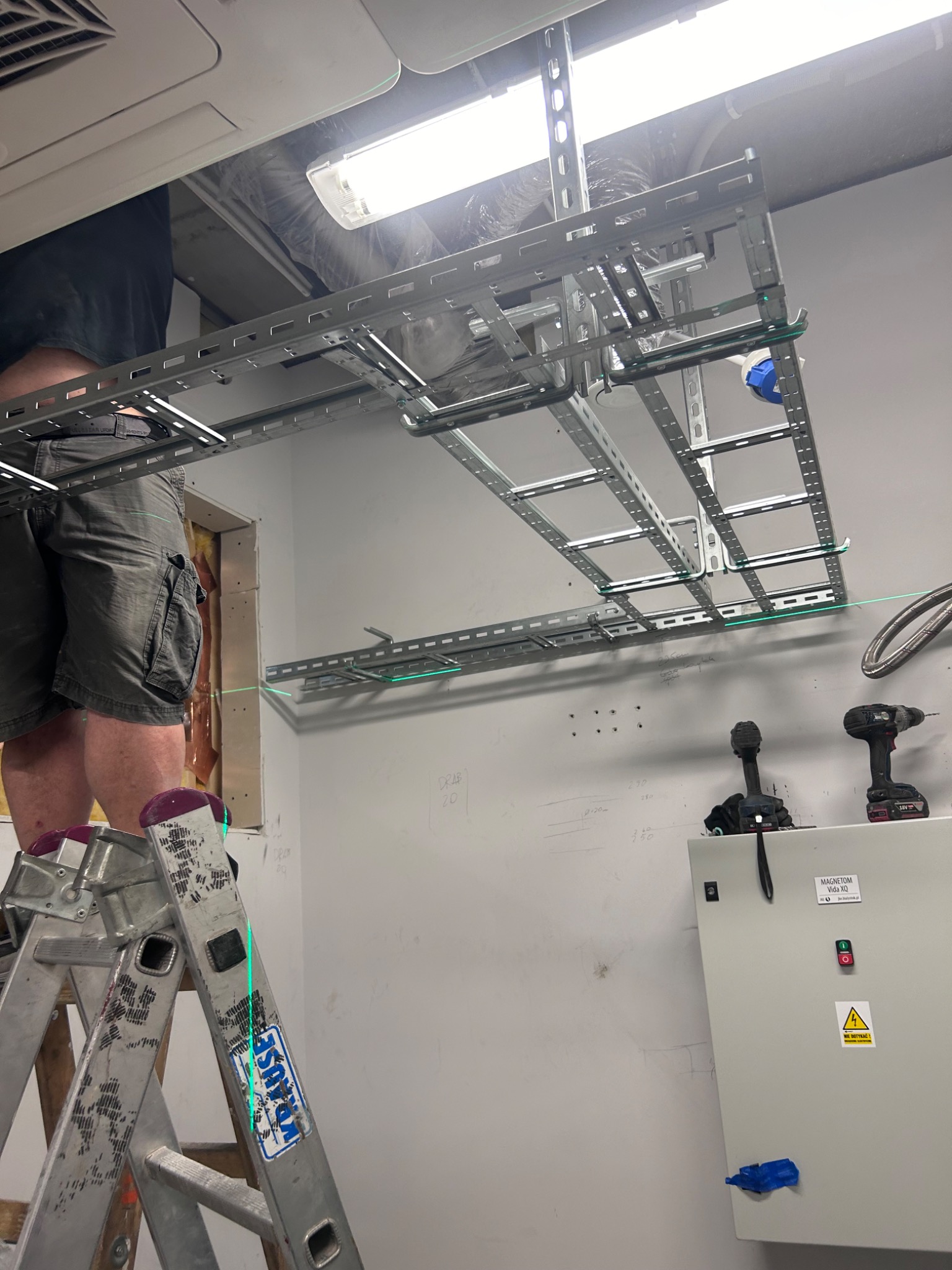 Instalacja metalowych korytek kablowych pod sufitem, widoczna drabina Krause, poziomica laserowa wyznaczająca linię, skrzynka elektryczna z ostrzeżeniem.