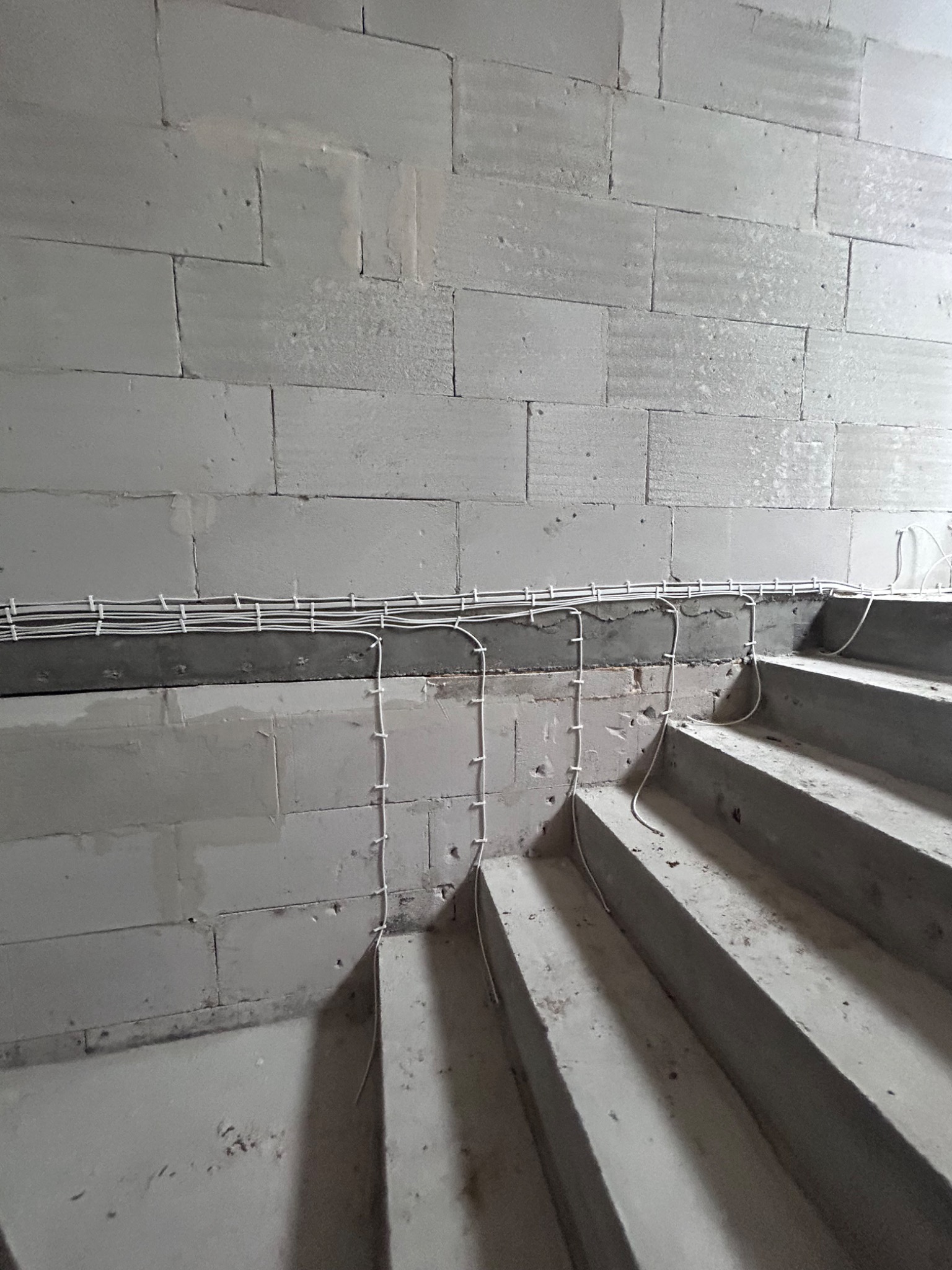 Surowa ściana z betonu komórkowego i betonowe schody z prowizorycznie poprowadzonymi przewodami elektrycznymi przytwierdzonymi do ściany i schodów za pomocą białych opasek zaciskowych.