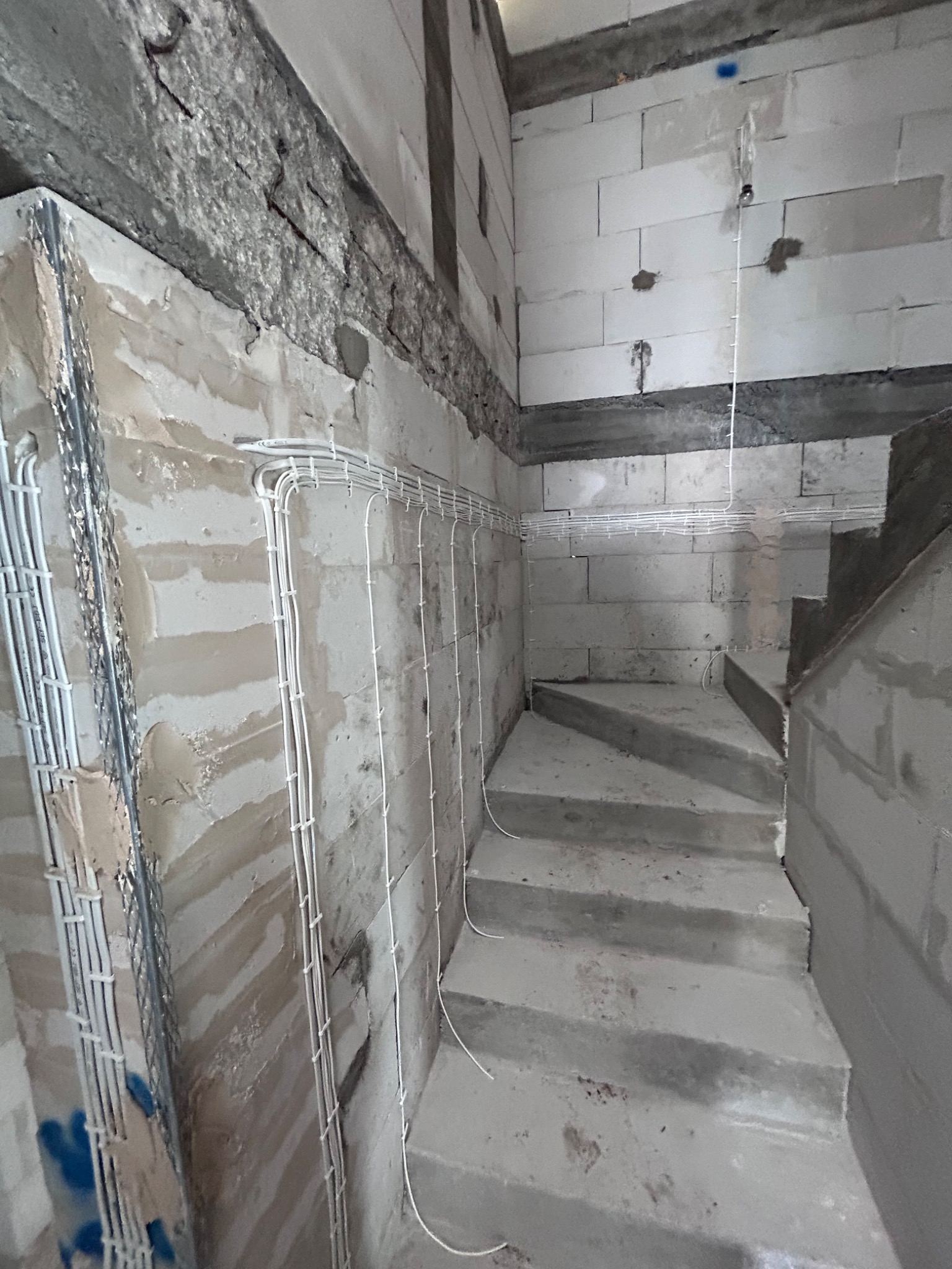 Wewnętrzne ściany budynku w trakcie prac wykończeniowych z widocznymi przewodami elektrycznymi poprowadzonymi w korytach i po ścianach, surowy beton schodów.