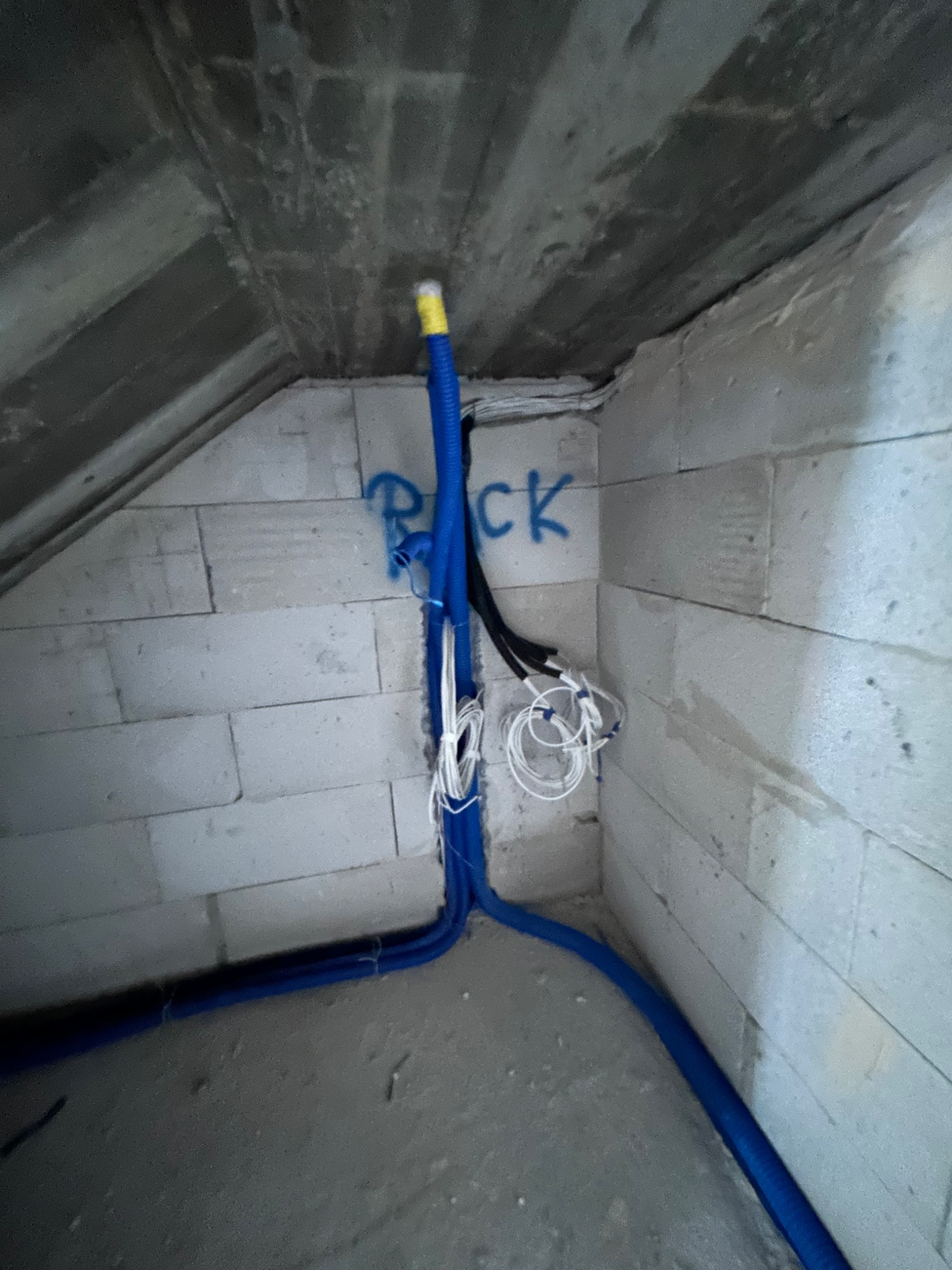 Niebieskie rury instalacji elektrycznej prowadzone po ścianie z betonu komórkowego w surowym wnętrzu poddasza.
