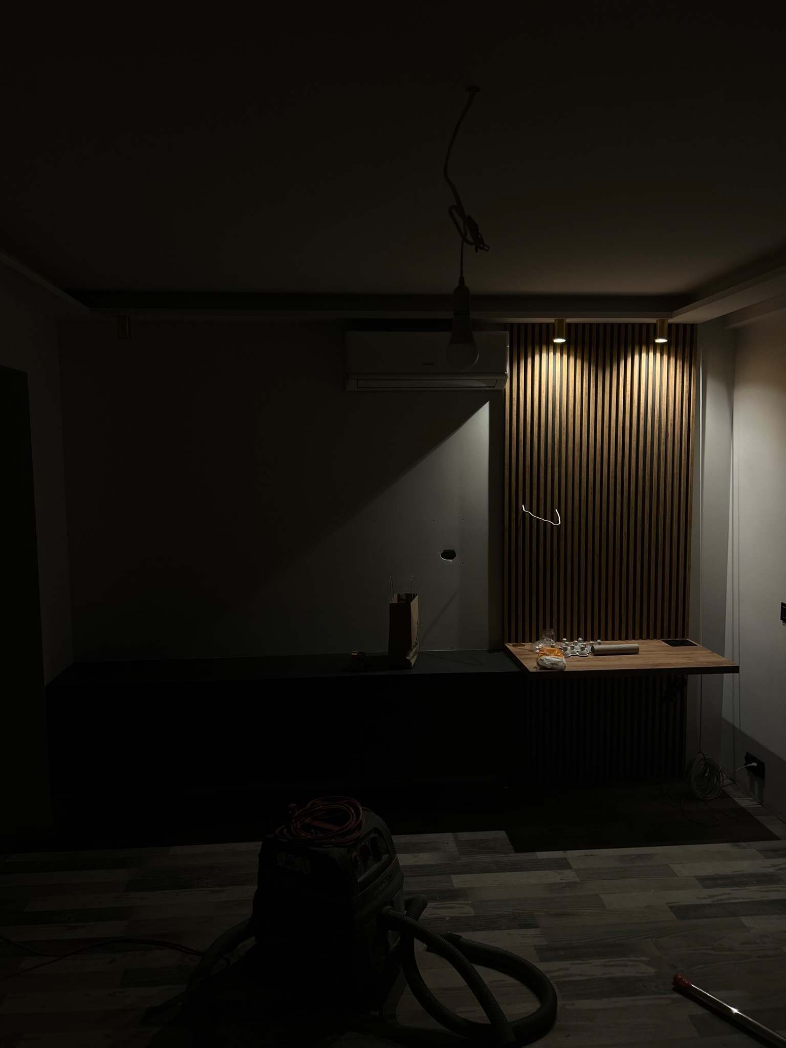 Fragment wnętrza w trakcie prac wykończeniowych: drewniana ściana z zamontowanym oświetleniem punktowym, widoczne przewody, narzędzia i odkurzacz przemysłowy na podłodze z imitacji drewna.