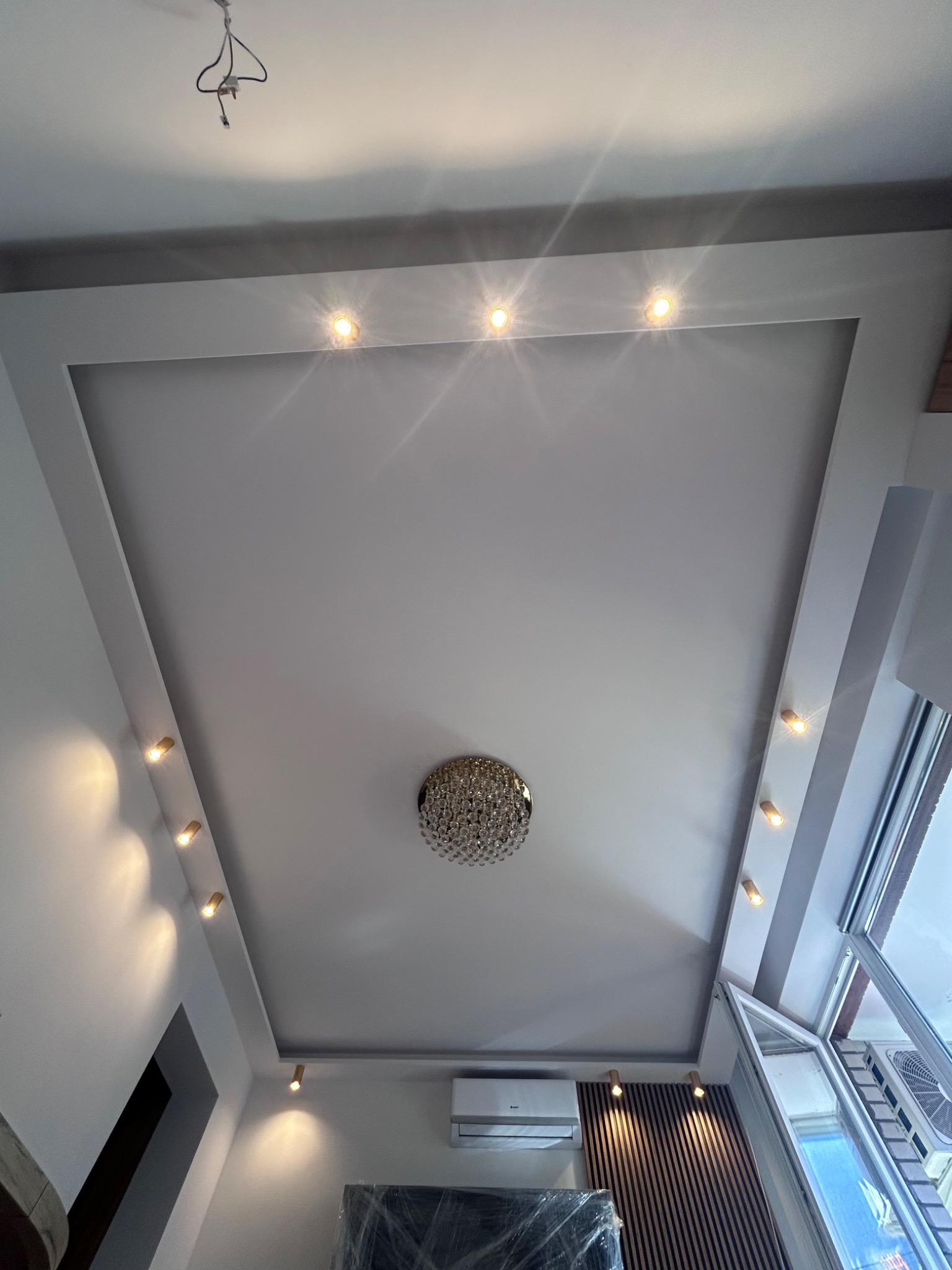 Nowoczesny sufit podwieszany z oświetleniem punktowym LED i kryształowym żyrandolem w centralnej części, widok z dołu, z widocznym klimatyzatorem i częściowo zasłoniętymi meblami folią ochronną.