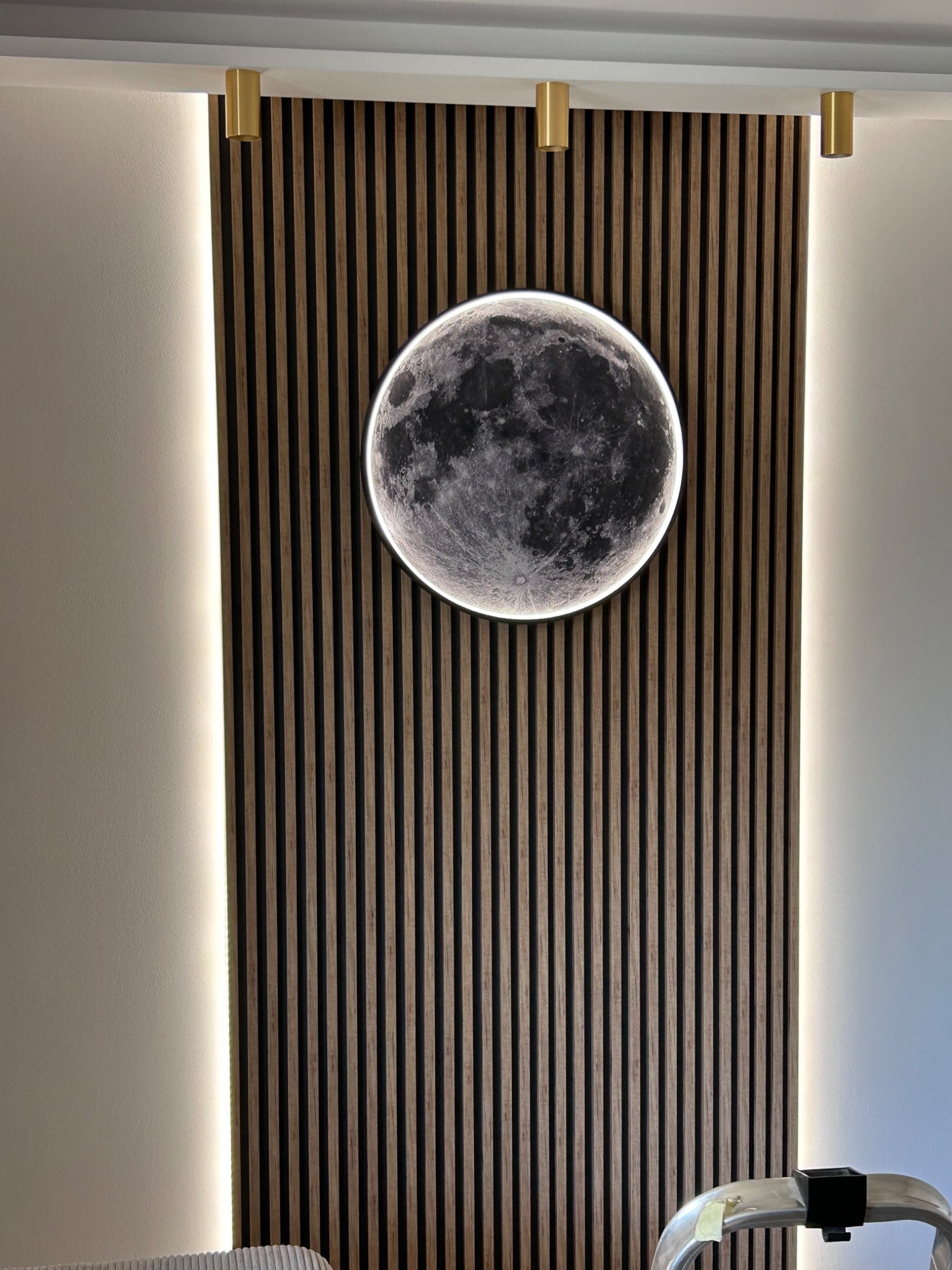 Okrągła lampa LED w kształcie księżyca zamontowana na drewnianej ściance z pionowych listew, oświetlona dodatkowo paskami LED po bokach, z widocznym fragmentem drabiny w dolnej części kadru.