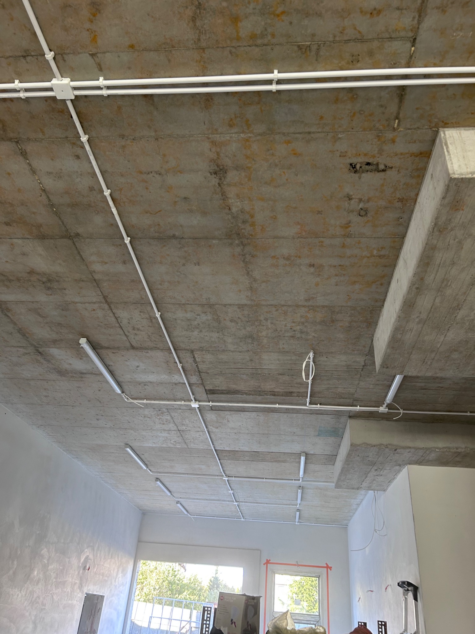 Surowy betonowy strop z widocznymi białymi rurami instalacji elektrycznej i przymocowanymi lampami liniowymi, widok z dołu.