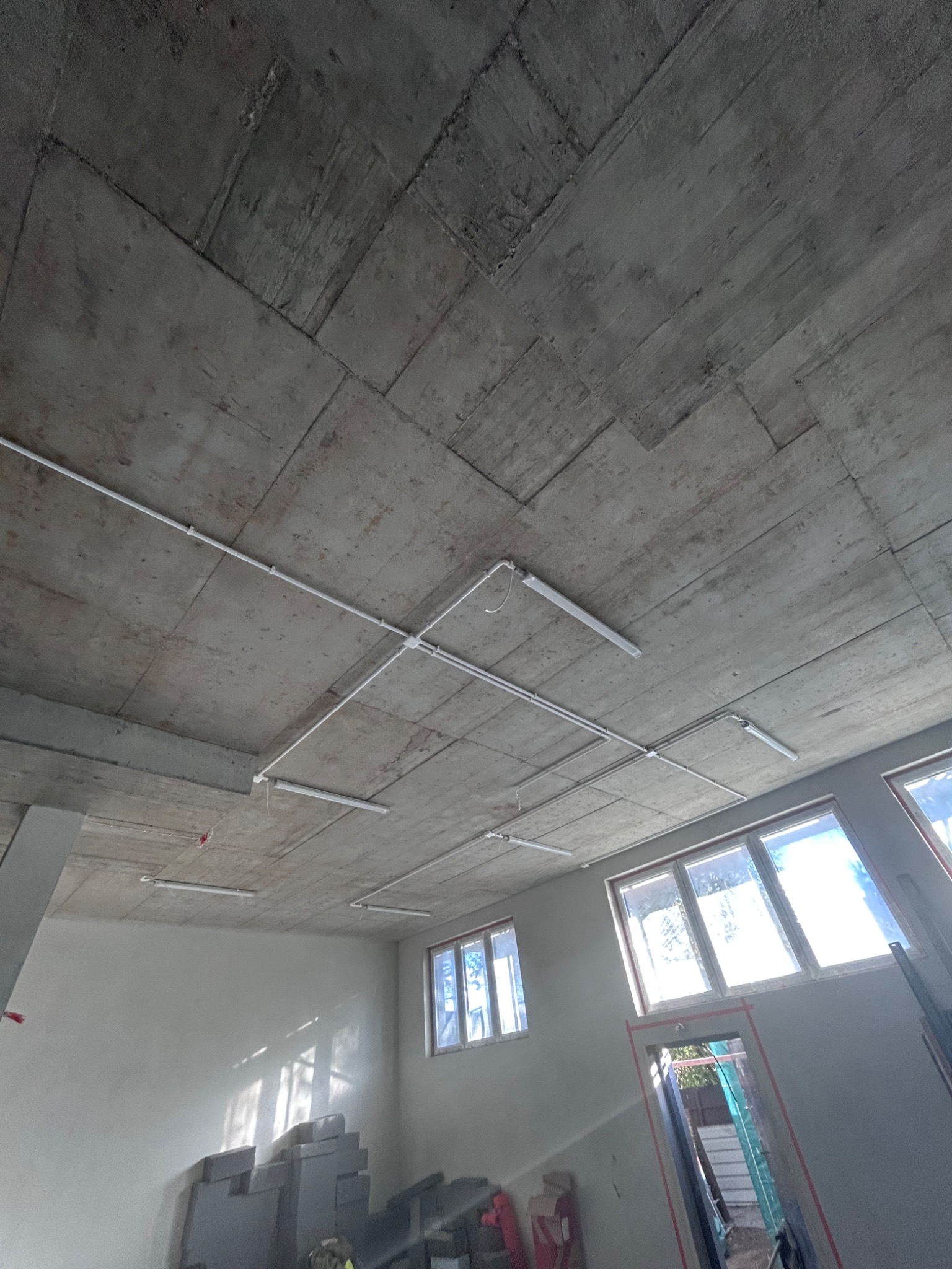 Surowy betonowy sufit z widocznymi przewodami i lampami fluorescencyjnymi, wnętrze w trakcie wykończenia, okna w tle.