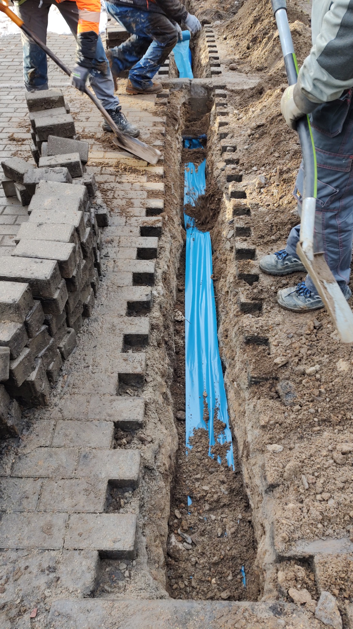 Wykop z niebieską folią ochronną na kable, obok stos kostki brukowej, robotnicy z łopatami zasypują wykop ziemią.