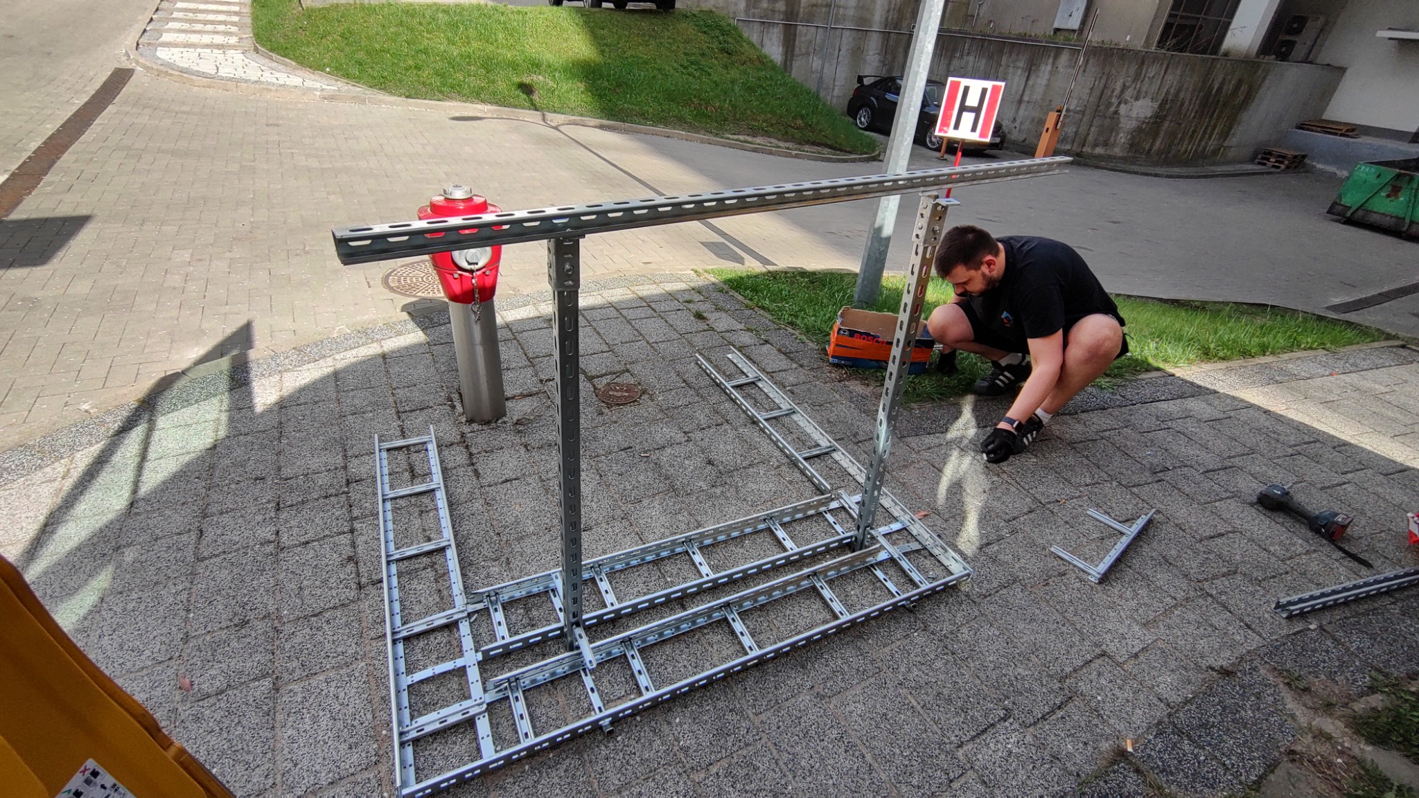 Pracownik w Białymstoku montuje metalową konstrukcję szkieletową na chodniku z kostki, obok czerwonego hydrantu i skrzynki narzędziowej Bosch, z użyciem wkrętarki akumulatorowej.