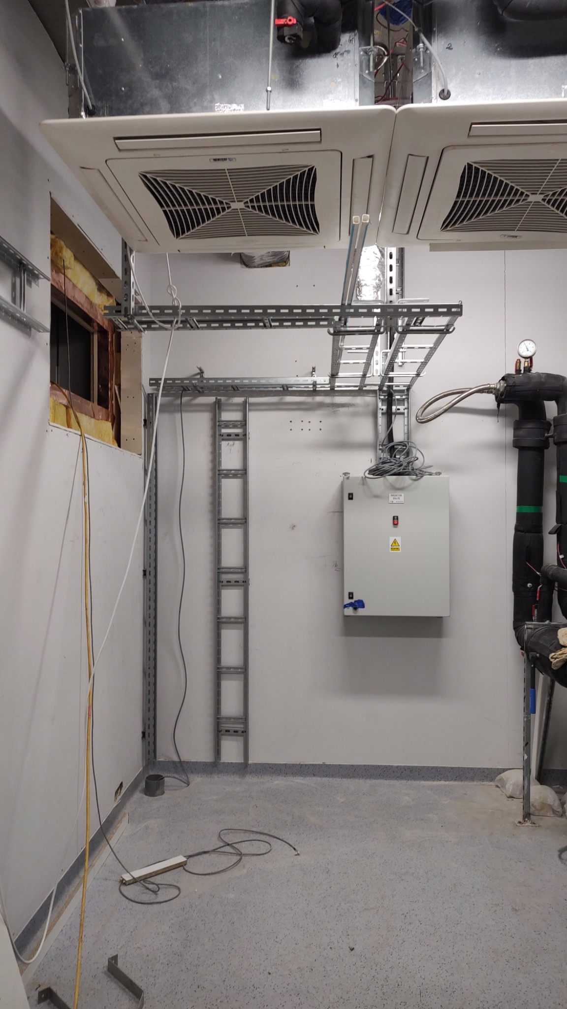 Pomieszczenie techniczne z zamontowanymi kanałami kablowymi, szafą sterowniczą i klimatyzatorami sufitowymi, widoczne rury instalacji grzewczej z manometrem oraz niedokończone prace przy ścianie...