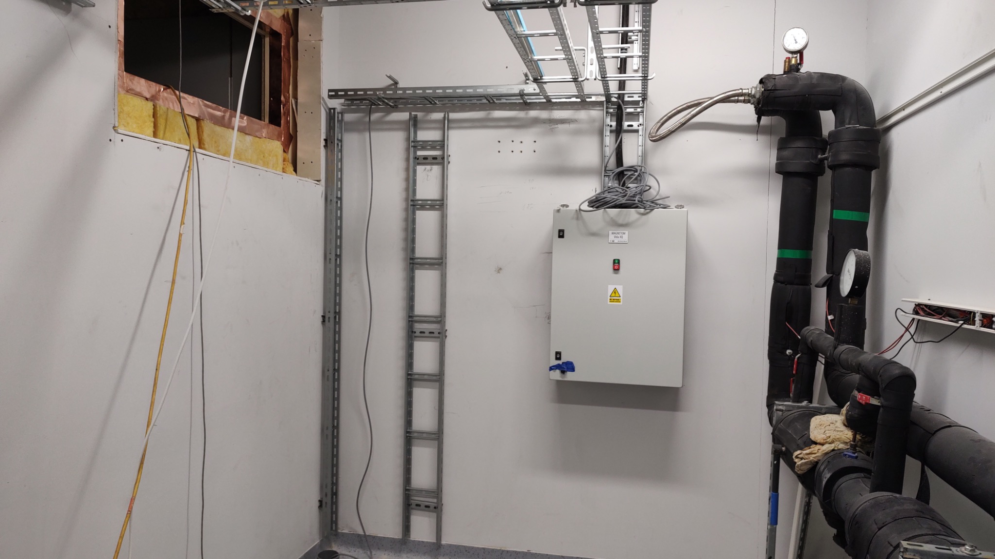 Wnętrze techniczne z metalowymi korytkami kablowymi pod sufitem, drabinką kablową na ścianie, szarą szafą sterowniczą z przyciskiem i znakiem ostrzegawczym, oraz izolowanymi rurami z manometrami.