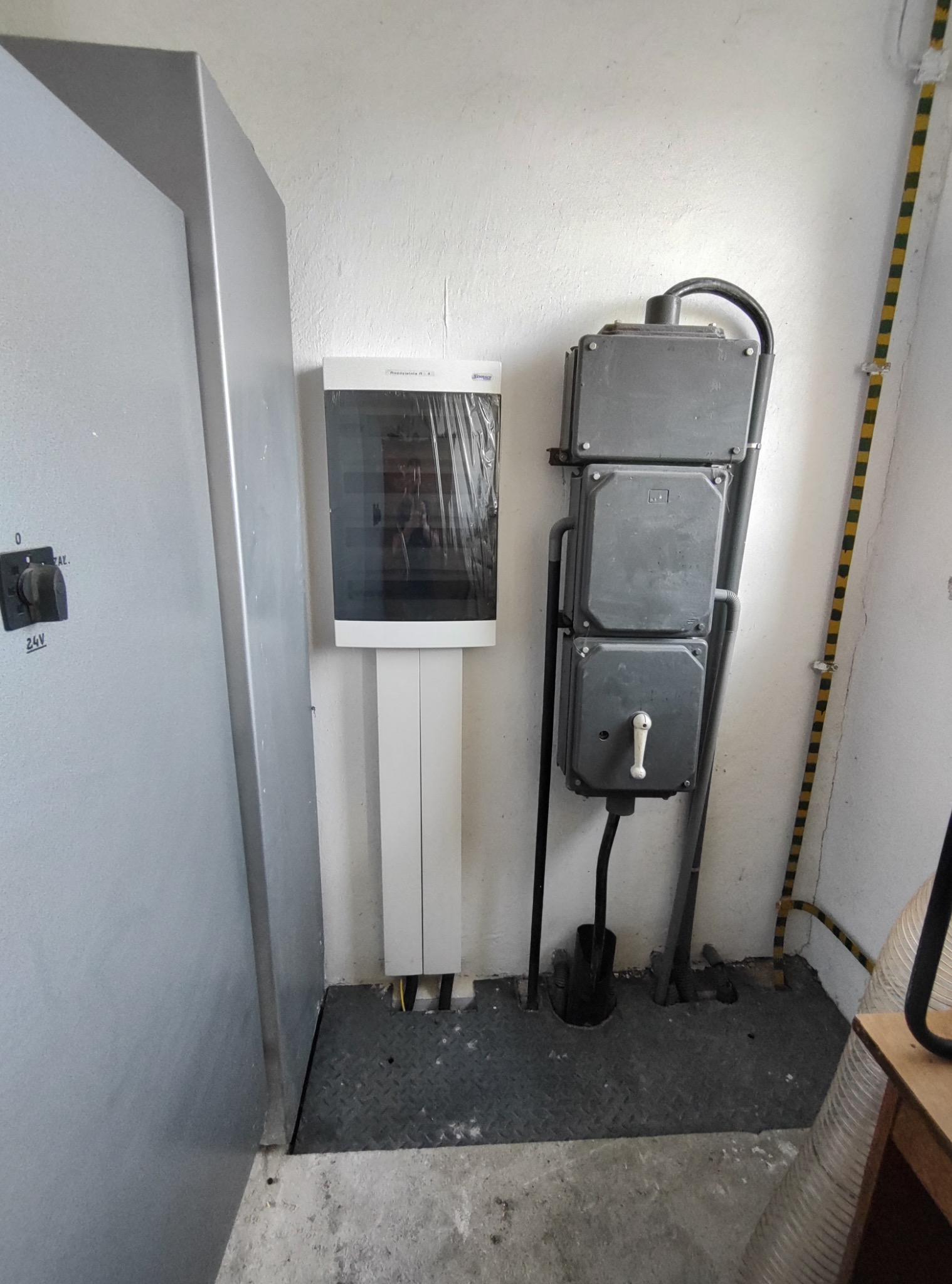 Szara szafa elektryczna z przełącznikiem, biała skrzynka elektryczna na ścianie i trzy szare puszki elektryczne połączone rurami, widoczne przewody elektryczne.