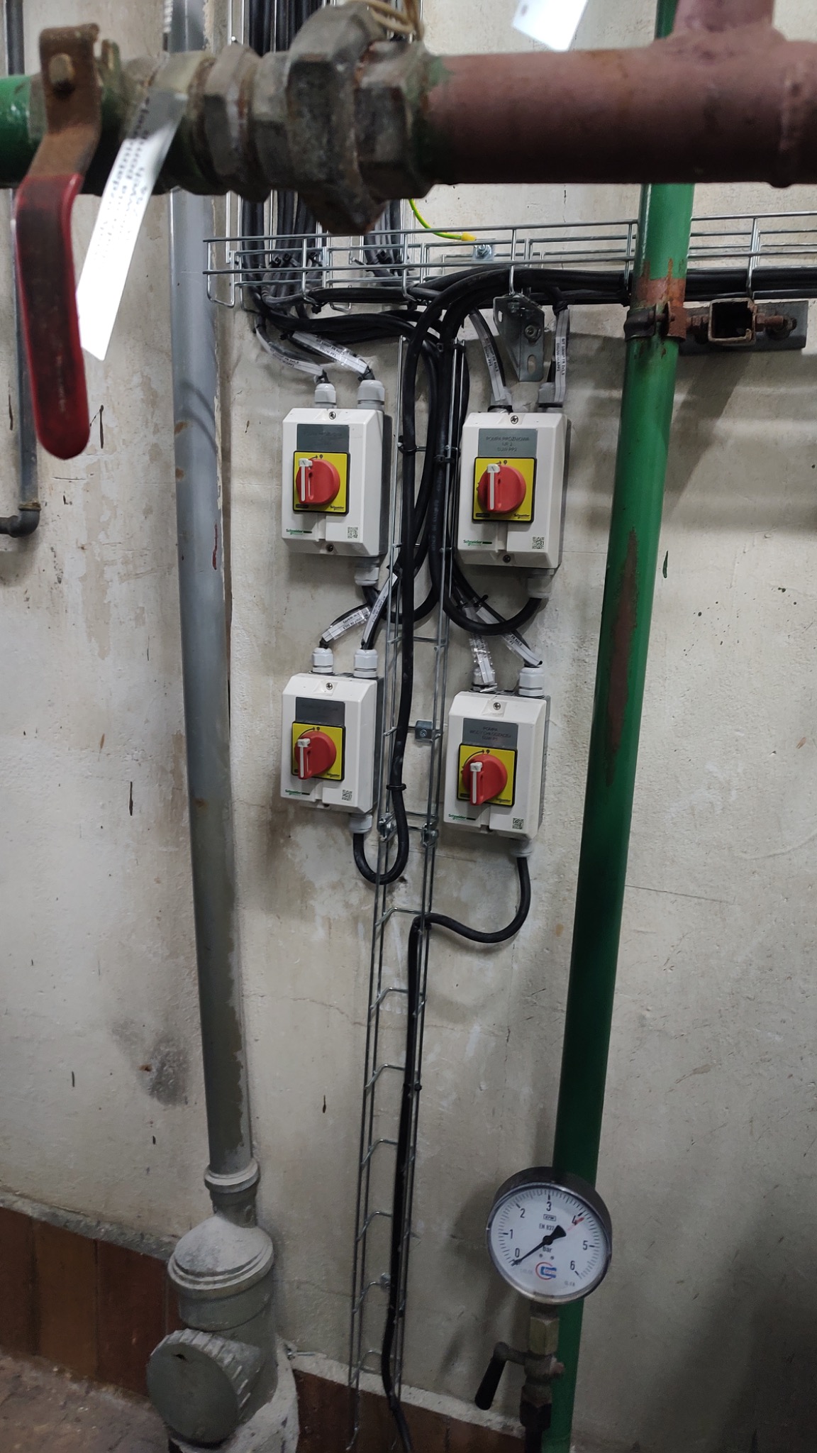 Cztery wyłączniki elektryczne z czerwonymi pokrętłami zamontowane na ścianie z widocznymi przewodami w metalowej korycie, obok rury wodociągowej i manometru.