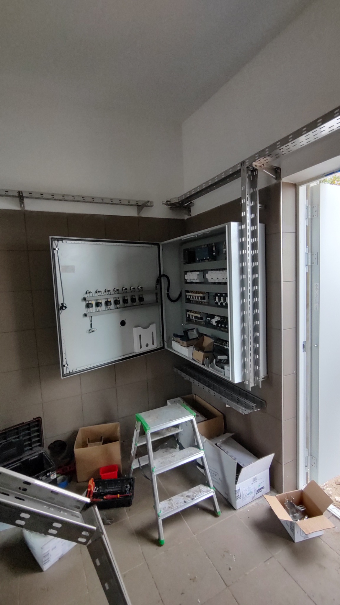 Otwarta szara szafa elektryczna z widocznym okablowaniem i elementami sterującymi, zamontowana na brązowych płytkach, drabina i narzędzia na podłodze.