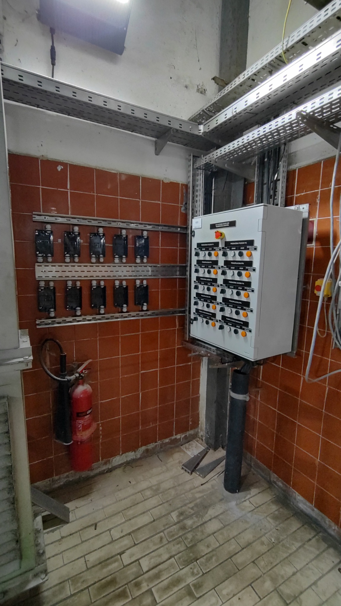 Zamknięta szafa sterownicza z przyciskami i lampkami, zamontowana w rogu pomieszczenia z brązowymi kafelkami, widoczne koryta kablowe i gaśnica.