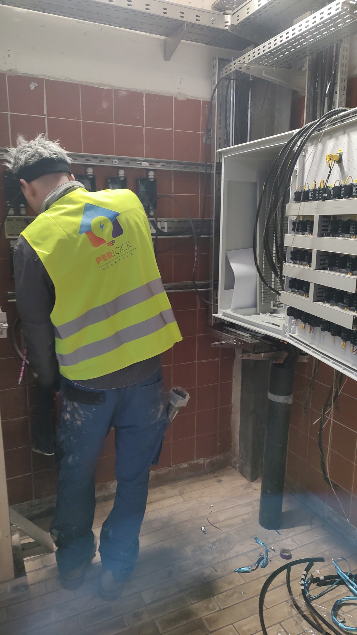 Elektryk w żółtej kamizelce z logo PerLock instaluje przewody w skrzynce rozdzielczej w pomieszczeniu z czerwonymi kafelkami, widoczne narzędzia i fragmenty przewodów na podłodze.