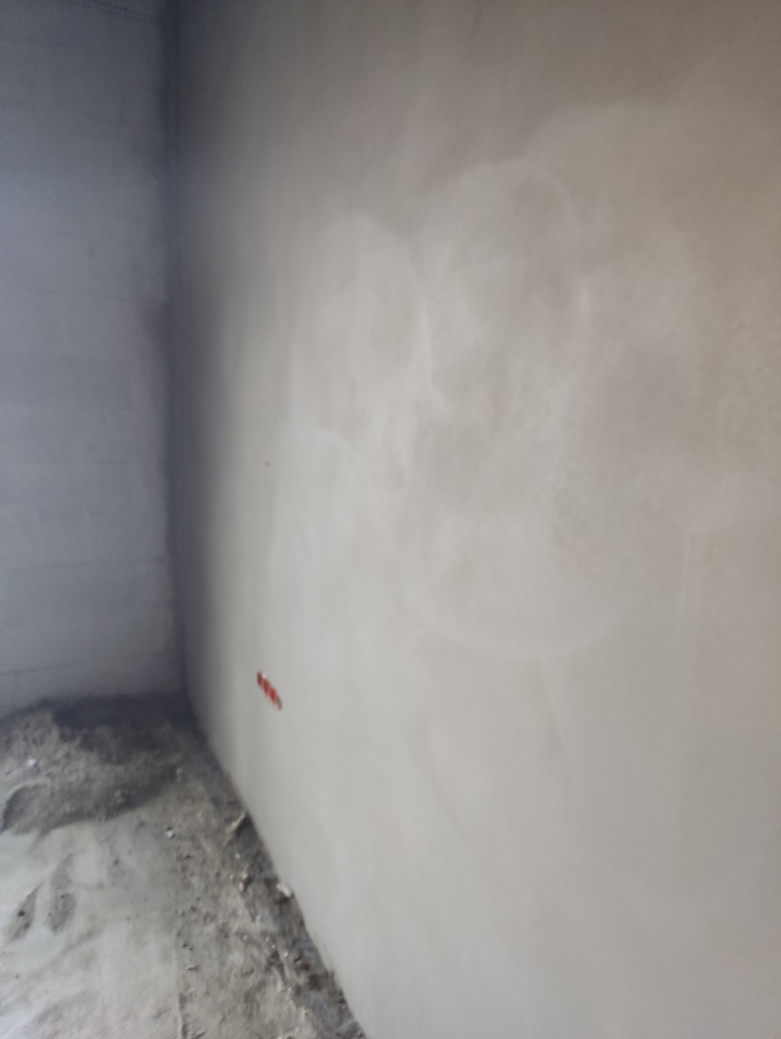 Świeżo otynkowana ściana w rogu pomieszczenia, widoczne ślady po łatach. Podłoga surowa, betonowa. Przygotowanie do dalszych prac wykończeniowych.
