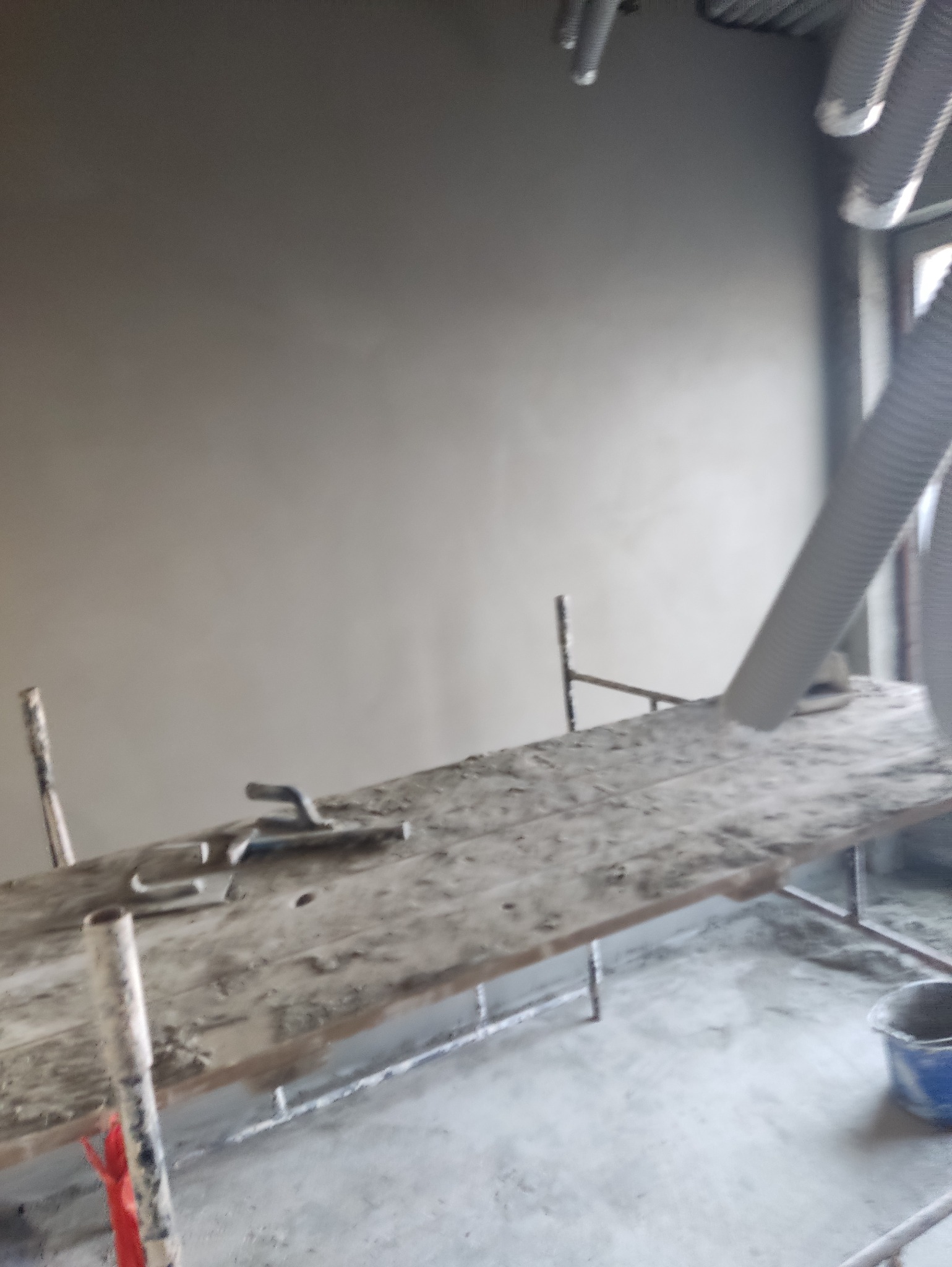 Świeżo otynkowana ściana w szarym odcieniu, widoczna z poziomu rusztowania z narzędziami. W tle rury wentylacyjne i okno.