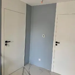 Dwa białe, nowoczesne drzwi wewnętrzne z czarnymi klamkami na tle szarej ściany i marmurowej podłogi, widoczne wystające przewody elektryczne na suficie.