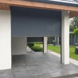 Ciemnoszara osłona przeciwsłoneczna częściowo opuszczona na tarasie, widok na zadbany ogród z meblami wypoczynkowymi w tle.