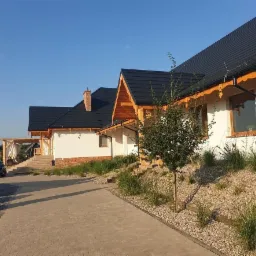 Elewacja domu jednorodzinnego z ciemną dachówką, drewnianymi elementami wykończeniowymi i ozdobnym ogrodem skalnym z trawami, widok z brukowanej drogi dojazdowej.