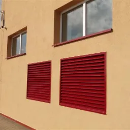 Elewacja budynku z dwoma oknami PCV i dwiema czerwonymi żaluzjami aluminiowymi wentylacyjnymi, widok z zewnątrz przy słonecznej pogodzie.