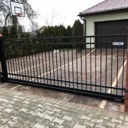Czarna, przesuwna brama wjazdowa z pionowymi prętami, zamontowana między ceglanymi słupkami na posesji z kostki brukowej, widoczny garaż i kosz do koszykówki.