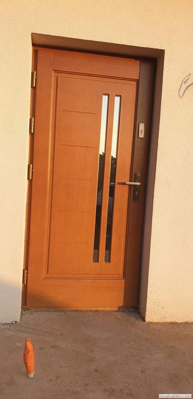 Brązowe drzwi wejściowe z dwoma pionowymi przeszkleniami, zamontowane w jasnej elewacji budynku, na dole widoczny pomarańczowy przedmiot.