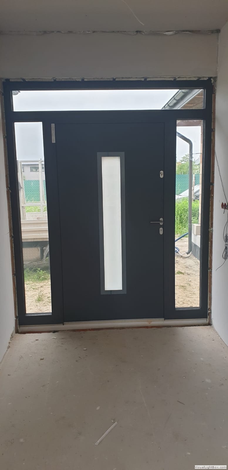 Nowoczesne, antracytowe drzwi wejściowe z przeszkleniem, zamontowane w nowym budynku w trakcie wykończenia, widoczne przewody elektryczne i surowy beton na podłodze.