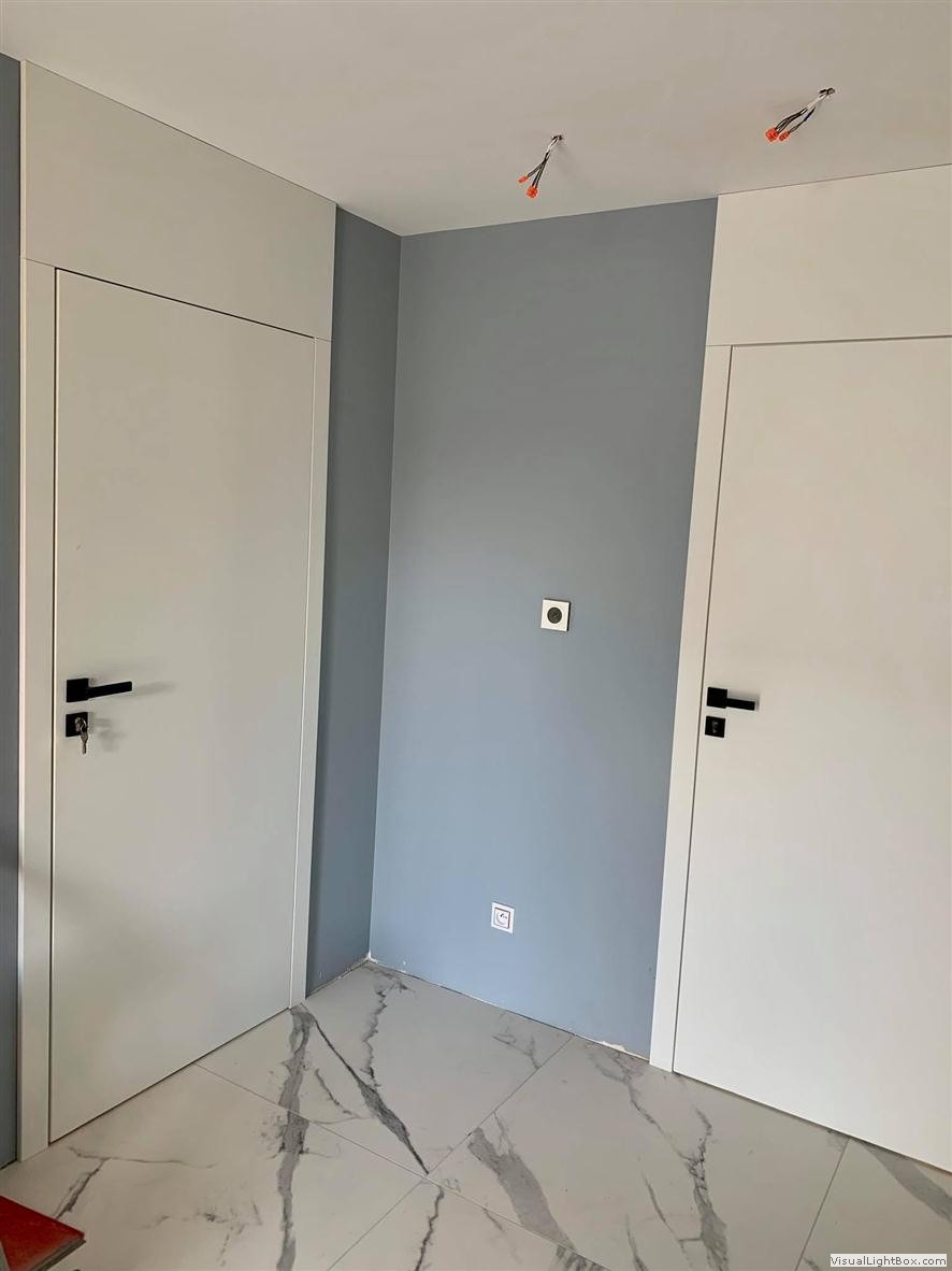Dwa białe, nowoczesne drzwi wewnętrzne z czarnymi klamkami na tle szarej ściany i marmurowej podłogi, widoczne wystające przewody elektryczne na suficie.