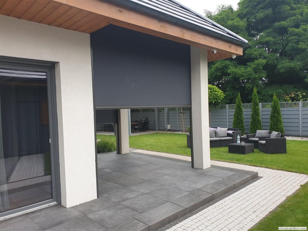 Zewnętrzna, grafitowa roleta zaciemniająca częściowo opuszczona, taras wyłożony szarymi płytami, w tle meble ogrodowe i trawnik.