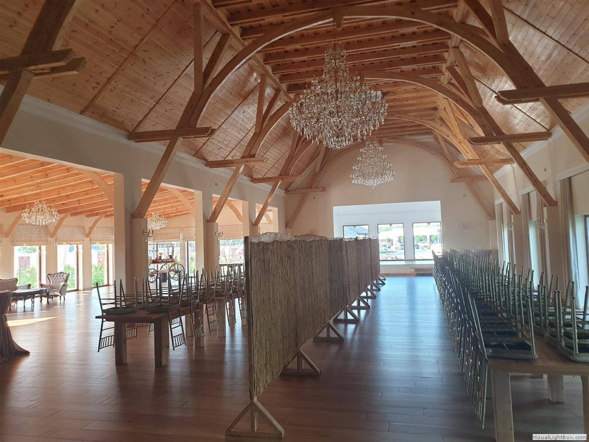 Duża sala bankietowa z drewnianym stropem, kryształowymi żyrandolami i podłogą z jasnego drewna, przygotowana do przyjęcia z krzesłami ułożonymi na stołach i parawanem z trzciny