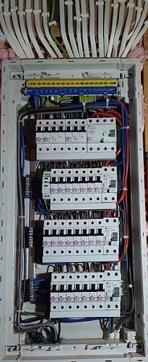 Rozdzielnica elektryczna z zabezpieczeniami, widoczne liczne przewody i bezpieczniki, zamontowana w ścianie z izolacją, ujęcie z góry.