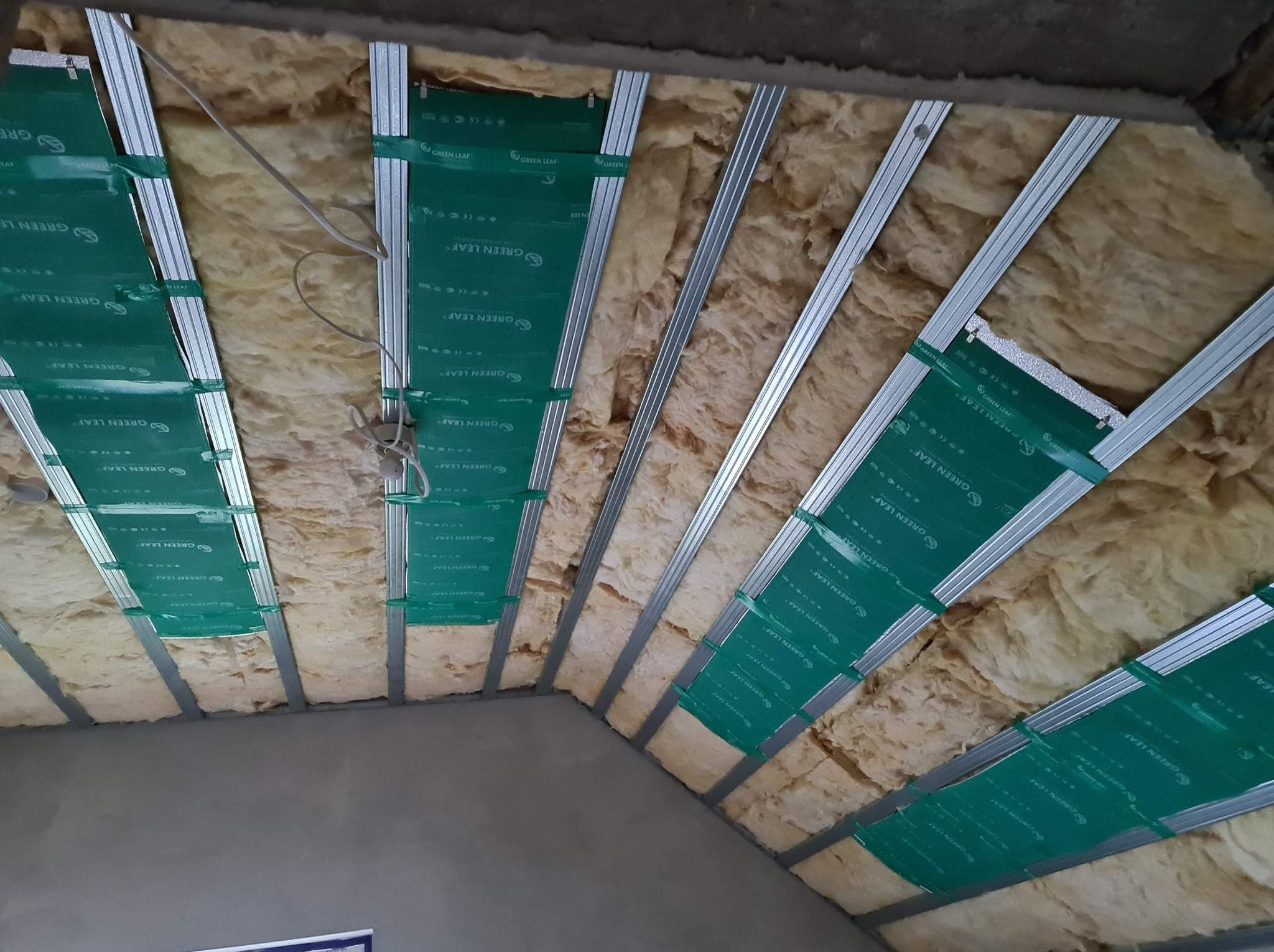 Ocieplenie poddasza wełną mineralną, widoczne metalowe profile i folia paroizolacyjna z nadrukiem 'GREEN LEAF', przewody elektryczne wystające z izolacji.