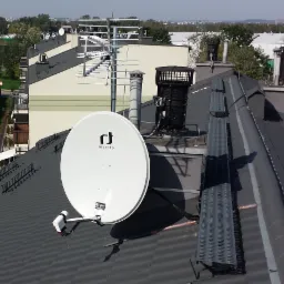 Antena satelitarna Invertro zamontowana na ciemnym, spadzistym dachu budynku wielorodzinnego, widok z góry na tle innych dachów, kominów i anten.