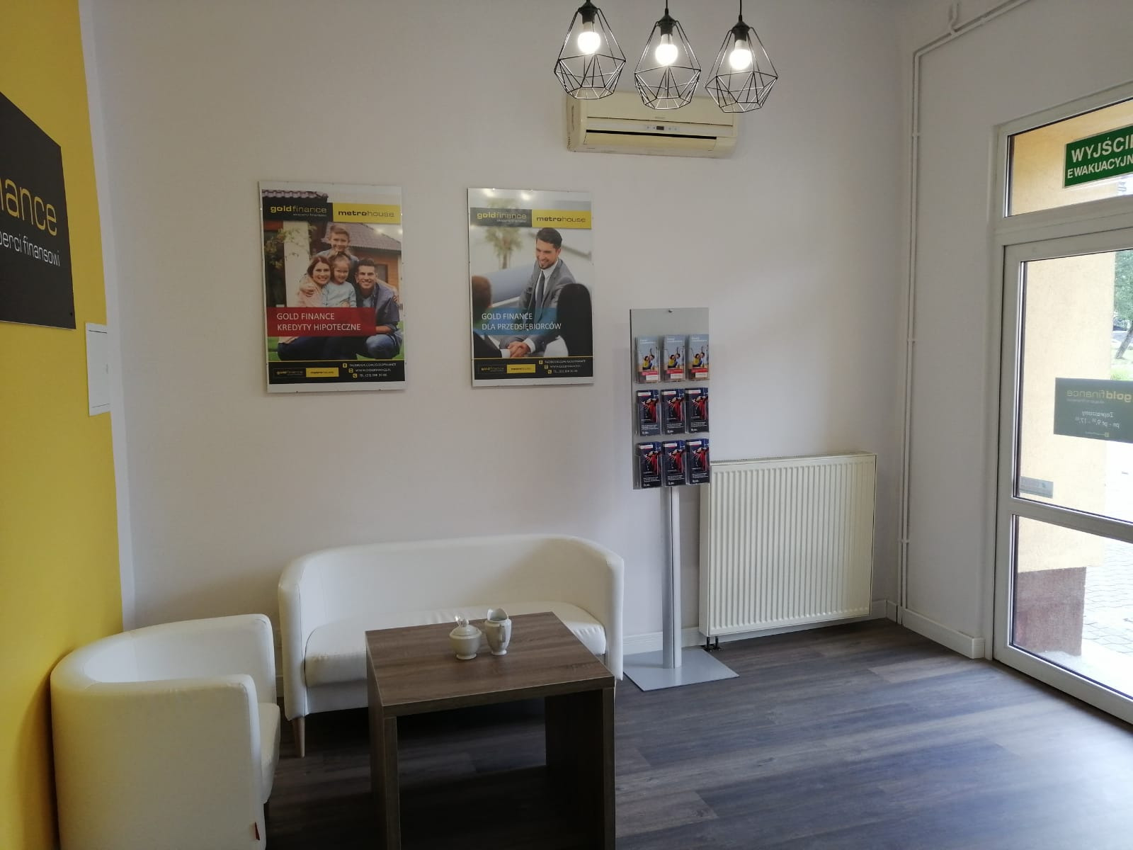 Poczekalnia biura finansowego z białymi meblami, stolikiem kawowym z cukiernicą i filiżanką, plakatami reklamowymi na ścianie, stojakiem z ulotkami i wiszącą klimatyzacją.