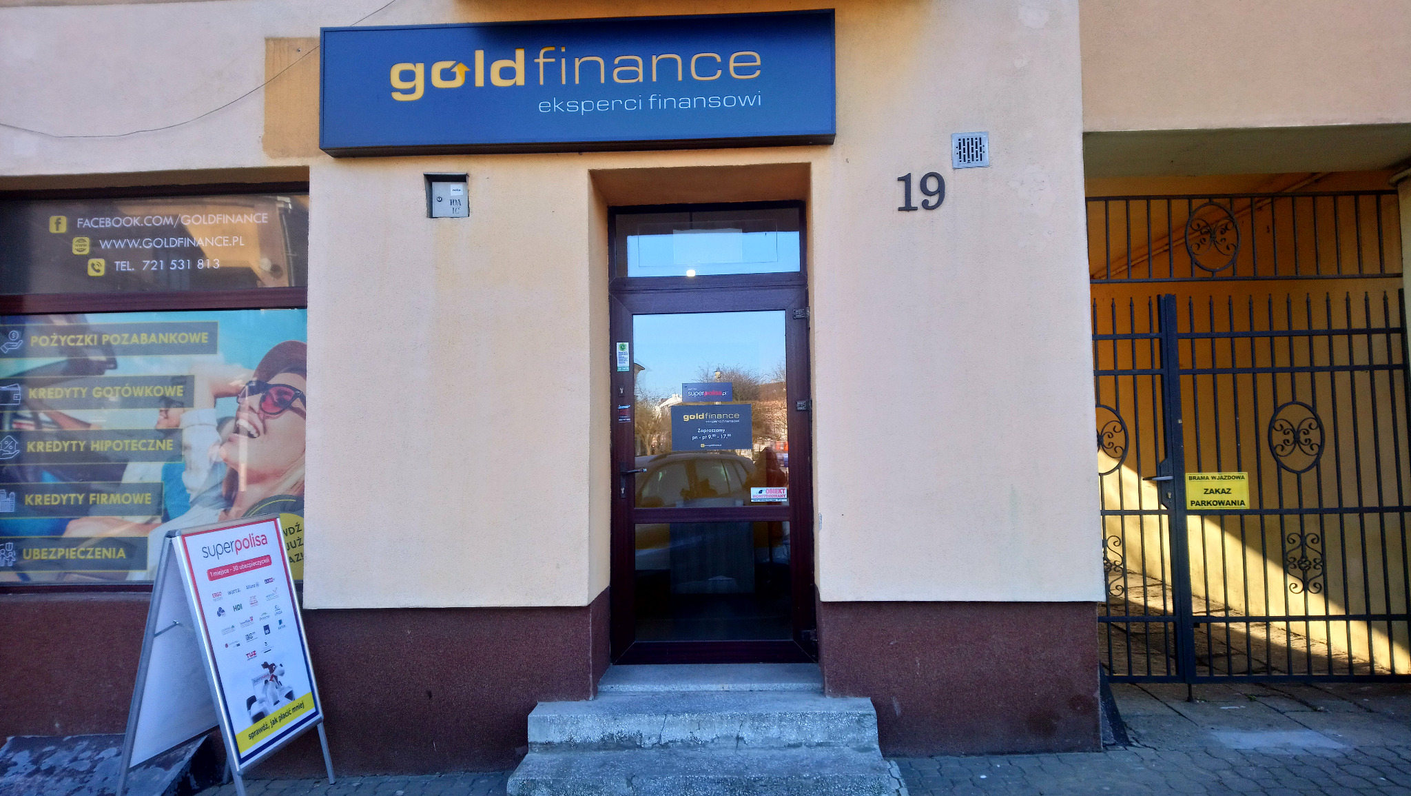 Fasada budynku z szyldem 'goldfinance eksperci finansowi', numerem 19, wejście z przeszklonymi drzwiami, obok reklama 'superpolisa' i brama wjazdowa z zakazem parkowania.